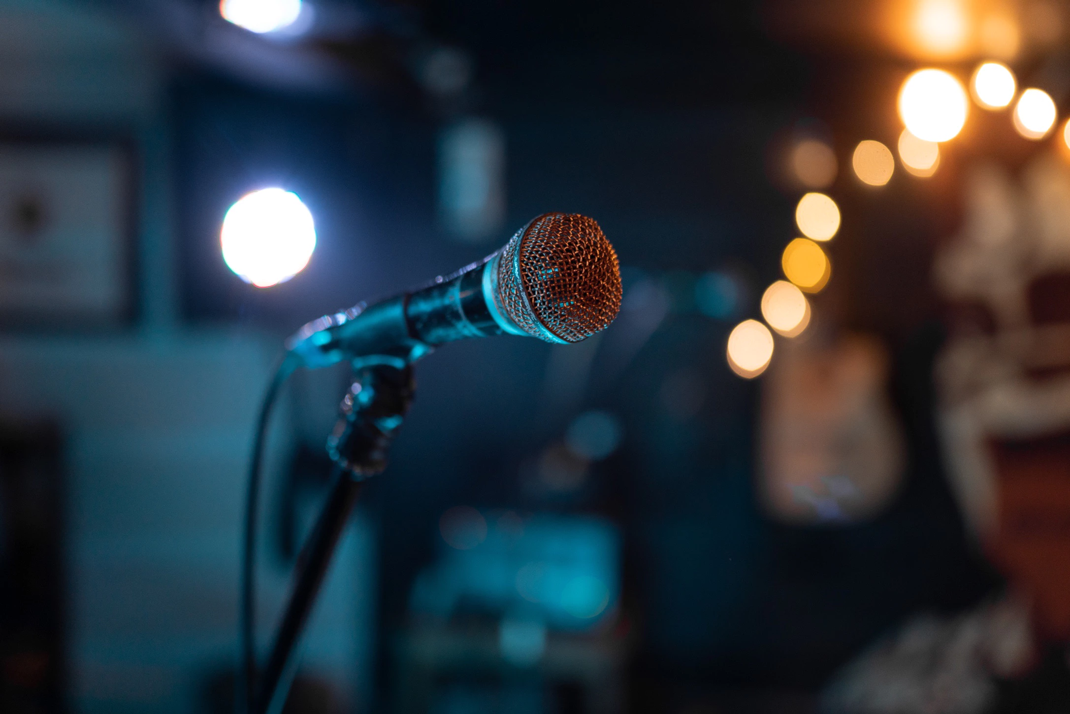 A close-up of a microphone on a stand in a dimly lit venue. The microphone's metal grille reflects warm light, while the background features blurred stage lights and musical instruments, creating a moody atmosphere.